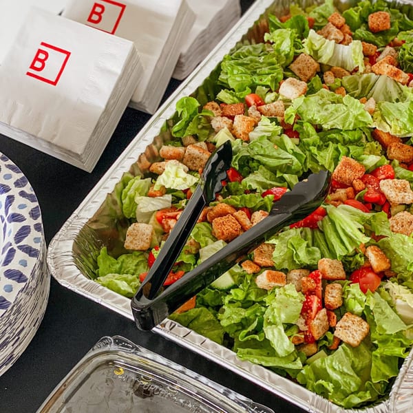A large catering tray of fresh green salad with croutons and tomatoes, with serving tongs.