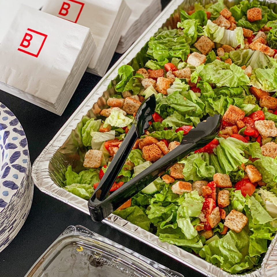A large catering tray of fresh green salad with croutons and tomatoes, with serving tongs.